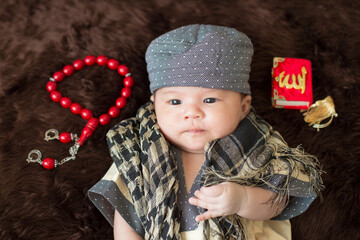 A cute Asian infant Muslim in thobe with prayer cap on bed in home.