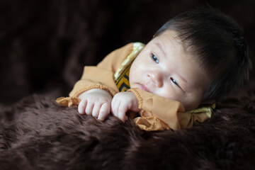 A newborn baby wearing a Chinese emperor's costume gold colour lying on a brown blanket.