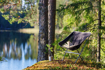 Camping folding chair with a book and a mug on the background of a forest lake. The chair is in focus, the background is blurred. - obrazy, fototapety, plakaty