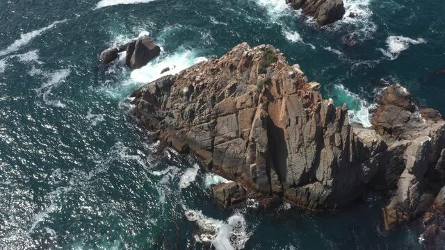 Aerial footage of a rocky cliff with waves crashing on it during a sunny day in Ensenada, Baja California, Mexico.