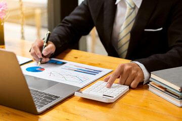 An accountant using a calculator and computer with a pen in hand is making money and analyzing graph papers on the table about expenses at home.