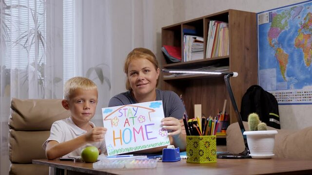 Portrait of an adult mother and son at home at a desk while doing homework, they hold a sheet with the inscription STAY AT HOME. Distance learning.