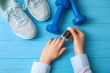 Female hands with pulse oximeter, dumbbells and sport shoes on color wooden background