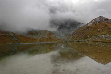 Mountain lake in autumn under low clouds, vegetation in autumn colors, moutains reflected in mirror-smooth water