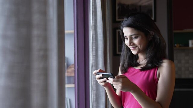Beautiful Indian girl holding smart phone watching funny videos,movie or social media content while standing near a widow in home office.Pretty woman looking at phone screen and watching funny content
