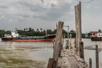 Fototapeta premium Bangkok, Thailand - 10 Jul 2020 : The old broken wooden pier by Chao Phraya River with cargo ship at the background.