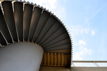 A gray spiral staircase around a thick pipe leads to a fork in the bridge or into a building. the railing is made of vertical sheets.