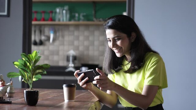 Asian Young Woman Play Mobile Games On Smart Phone And Win With Fist Gesture At Home. Smiling Indian Woman Having Fun And Enjoying In Free Time.Attractive Asian Woman Playing Game On Smart Phone.