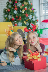 Two young caucasian girls with blonde hair wearing Christmas theme sweaters celebrating and enjoy Christmas at home with happiness. Christmas trees in the background