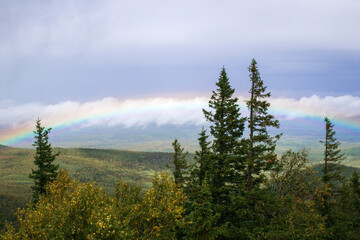 View on mountains landscape with fir trees on foreground and cloudy sky with rainbow on background. Ural Mountains, Russia.