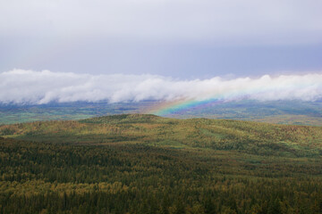 Scenic landscape with mountains and forest and cloudy sky with rainbow on background. Ural Mountains, Russia.