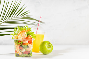 Healthy fitness food - set of fresh orange juice, green apple, tropical salad with shrimps and vegetables in plastic box in white interior with green palm leaf. Organic cuisine takeaway.