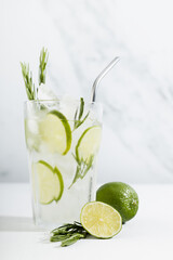 Organic mojito cocktail with citrus fruit slices, ice cubes, rosemary twig, silver straw, ingredients in transparent glass on white wood table, soft light marble wall, vertical.