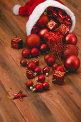Close up shot of red decorative hanging items star shiny glossy sphere balls ribbon bow tie and present gift boxes pouring from Santa claus Christmas hat on dark brown wooden table with copy space