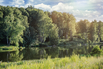 A pond with banks covered with lush grass and trees against a bright cloudy sky with white clouds and light leaks. Rural landscape in summer.