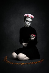 Painted Catrina woman and flower crown on her knees with a flower in her hand on dark background,...