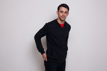 Smiling young brunette man in dark blue jumper and jeans posing near white wall background.