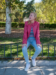 Girl wearing jeans red leather jacket and white sneakers sitting on park railing with trees and grass behind her