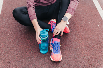 Sportswoman in stylish clothes and sneakers with bottle of water and smartwatch sits resting on track at stadium close view