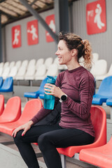 Happy curly haired lady in comfortable tracksuit with bottle of water and smartwatch sits resting on plastic chair at stadium