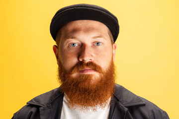 Close-up portrait of stylish red-bearded man, barber in black cap looking at camera isolated over yellow background. Concept of emotions, feelings.