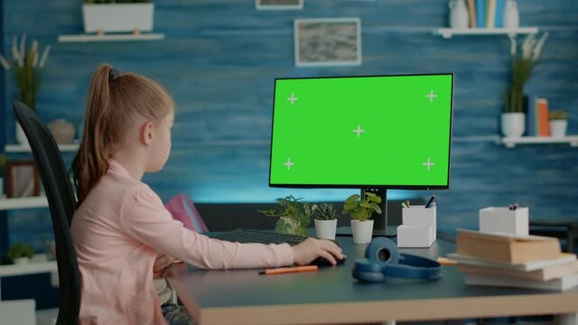 Schoolgirl looking at computer with horizontal green screen at home for remote courses and classes. Young child using monitor with chroma key and isolated mockup template at desk.