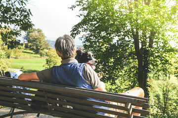 Fototapeta premium Man and his dog sitting on a park bench looking the mountain panorama. Friends together relaxing at the park. Guy with pet outdoor in the nature. Friendship, loyalty family and companionship concept