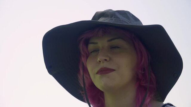 Mujer de pelo rosa con sombrero  grande y labios rojos, Mujer con sombrero mirando a los lados y sonriendo , chica joven con sombrero y de fondo el cielo blanco y nublado