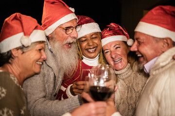 Diverse senior people having fun cheering with wine during Christmas eve wearing Santa Claus hats - Soft focus on african woman face