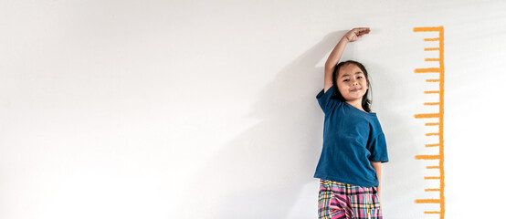 An adorable Asian little child girl measuring the height growth with the orange drawing on the white wall background. Girl power future and dream concept idea. Copy space - obrazy, fototapety, plakaty