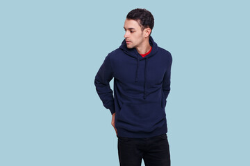 Profile of a young man in dark blue jumper and jeans, holding his hands in his pockets, isolated light blue background.