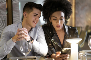 interracial young couple sitting at the restaurant table choosing food on digital menu thru the smartphone