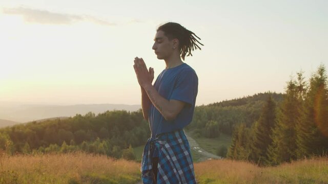 Peaceful handsome hipster arab man meditating in prayer yoga pose , clasping hands in namaste gesture, experiencing spiritual and mental healing while standing on mountain top at daybreak.