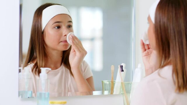 beauty, hygiene and people concept - teenage girl looking in mirror and cleaning face skin with cotton disc at bathroom