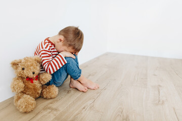 Lonely scared sad little boy with teddy bear sitting on floor in empty room