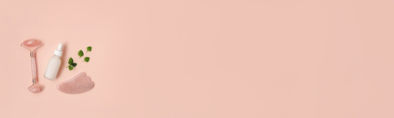 Composition of white mockup serum bottle, pink quarts stone face roller and gua sha stone with fresh leaves on pink background. Natural cosmetics concept. Creative cosmetics web banner flat lay with