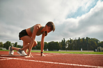 Concentrated young female athlete getting ready to start running on track field in the daytime