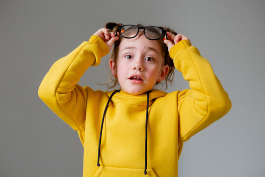 Portrait Of A Funny Cute Little Girl In Black-rimmed Glasses In A Yellow Hoodie With A Funny Hairstyl On A Gray Background. Much Studying. Child Who Is Tired Of School Activities. High Quality Photo