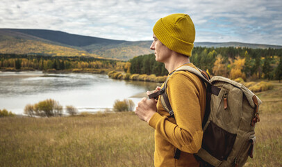 Man in a yellow hat and sweater with a backpack is standing in front of the river, autumn golden forest and hills