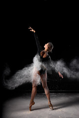 Fototapeta na wymiar Art perfomance. Full-length portrait of talented flexible ballet dancer moving in cloud of dust, young dancer in flour. Isolated over black studio background. Dance, people, art, performance concept