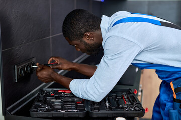 Side view on Electrician repairing sockets of residential electrical system,using screwdriver tool. Skilled confident african Handyman in blue overalls during work, concentrated and focused