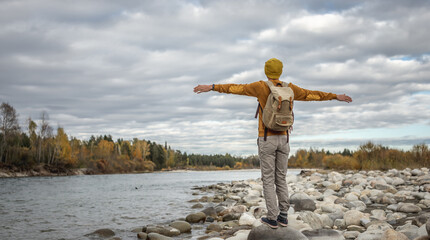 Traveler is standing with his arms outstretched to the sides next to fast river with big stones on the shore