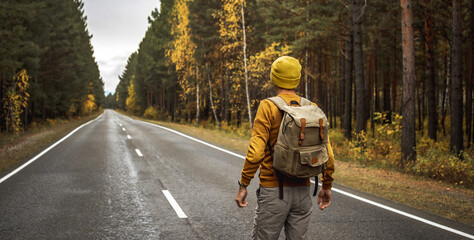 Traveler with a backpack is walking on an empty road along forest. Concept of freedom, travel,...