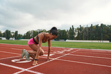Professional young female runner in sportswear getting ready to start running on track field at...