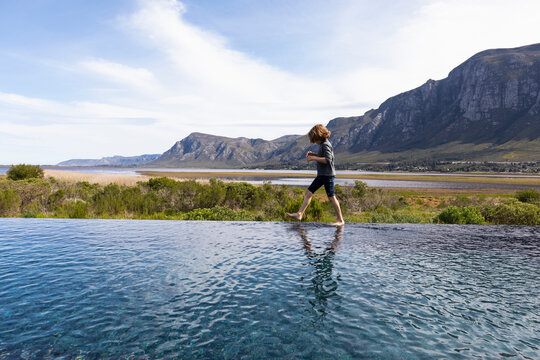 Eight year old boy walking around the edge of an infinity pool, a mountain backdrop