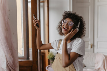 close-up of cheerful well-groomed teenager enthusiastically listening to favorite song on headphones. young african woman with closed eyes holds black mobile phone in bright room.