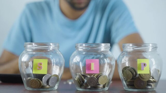 Focus on jar, Man from behind placing coins inside the jar - Concept of monthly SIP or systematic investment plan.