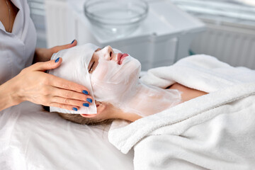 people, beauty, spa, cosmetology and skincare concept. beautiful young woman lying with closed eyes while cosmetologist applying facial mask and cloth mask in spa cosmetology room. copy space