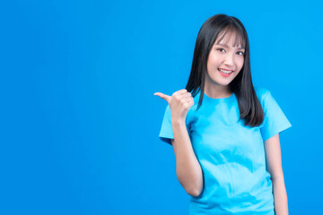 Fototapeta premium Beautiful Asian woman cute girl with bangs hair style in blue t shirt smiling and pointing finger to empty copy space for present product or blank space for advertising isolated on blue background