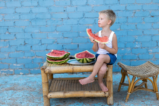 Cute boy in white T-shirt sits at wooden table in yard and eats watermelon. All hands and clothes are stained with watermelon juice